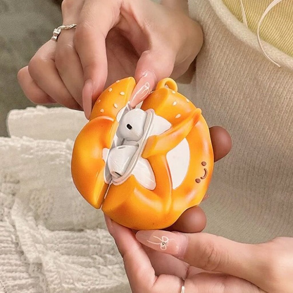 AirPods case designed to look like a smiling cartoon pretzel with a light brown silicone finish. The case is made of soft, textured material and includes a fun 3D design that covers the entire AirPods case.